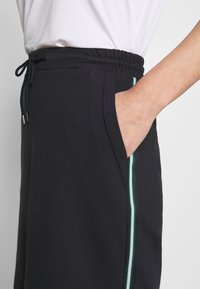 Pantaloni sportivi neri con vita elasticizzata e coulisse. Presentano tasche laterali e strisce verticali verdi e bianche lungo i fianchi.