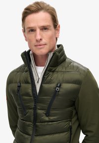 Padded jacket w kolorze oliwkowym z zamykanymi na zamek kieszeniami, czarnymi akcentami, wysokim kołnierzem i lekką teksturą. Warstwowa z szarym wewnętrznym podszyciem.