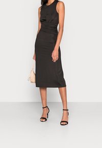 Schwarzes ärmelloses Kleid mit taillierter Silhouette, geraffter Taillendetail und knielangem Saum. Accessoires sind eine kleine beige Handtasche und schwarze Sandalen.