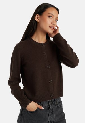 Cardigan marrone lavorato a maglia con collo rotondo, dotato di chiusura a bottoni, polsini a costine e design corto. Abbinato a jeans scuri.