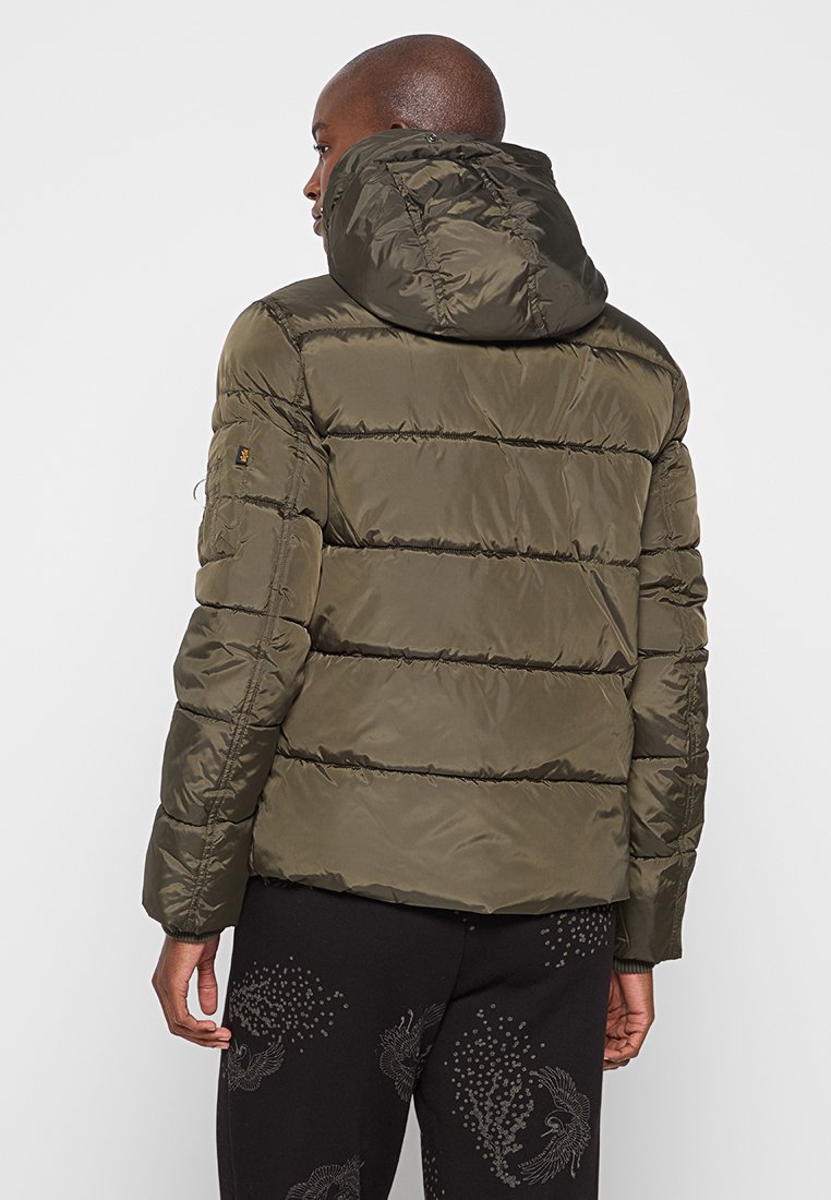 Hombre de espaldas vestido con una chaqueta de plumas verde oliva con capucha y pantalones negros con ligeros patrones decorativos.