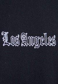 Melnā auduma gabals ar baltu izšūtu tekstu, kas lasāms "Los Angeles" gotiskā stilā ar grezniem elementiem un mainīgu līniju biezumu.