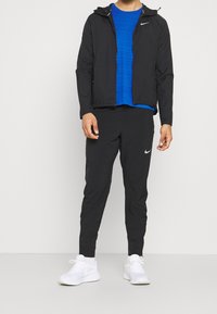 Černá lehká bunda s kapucí, modré texturované tričko, černé zúžené kalhoty a bílé sportovní boty s brandingem Nike.