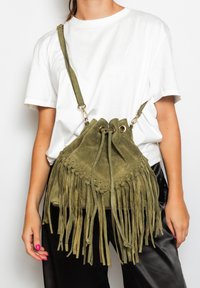Borsa a secchiello in camoscio verde oliva con frange, chiusura con coulisse e dettagli metallici; indossata a tracolla con spallaccio regolabile.