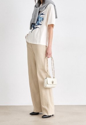 Pantalon beige à jambes larges, t-shirt crème à motif graphique bleu, pull gris noué autour du cou, talons noirs et petit sac à main blanc avec une bandoulière chaîne.