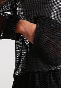 Nära bild av en svart mesh-tröja med långa ärmar. En hand är placerad i fickan, vilket framhäver det texturerade tyget och designen.