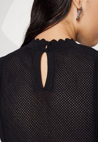 Černý pletený top s vlnitým výstřihem, s klíčovou dírkou na zadní straně a dvěma knoflíky na zapínání. Texturovaný, prodyšný design.