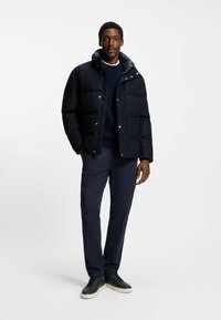 Giacca puffer blu navy con colletto alto e bottoni a pressione, abbinata a un maglione scuro, pantaloni navy e sneakers nere. Texture liscia.