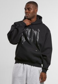 Black hoodie with a large glossy graphic on the chest, kangaroo pocket, and ribbed cuffs. Worn with gray sweatpants.