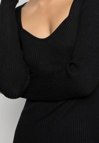 Top in maglina nera con costine, caratterizzato da un scollo a barchetta e maniche lunghe con polsini arricciati, che mette in risalto la texture e la silhouette aderente.