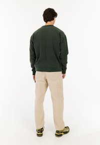 Donkergroene sweatshirt van zacht materiaal, gecombineerd met beige cargobroek met zijzakken. De outfit wordt aangevuld met donkere schoenen.