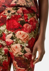 Floral pants made from soft fabric, featuring a vibrant red, pink, and green rose pattern. Includes front pocket and button closure detail.