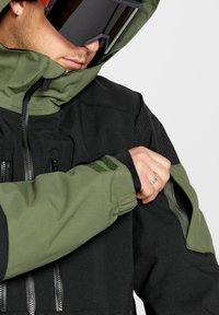 Giacca impermeabile nera e verde con una texture liscia, tasche con zip, polsini regolabili e una ventola con zip sulla manica.
