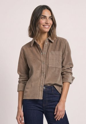 Femme aux cheveux mi-longs portant une chemise marron en velours côtelé à boutons et un jean bleu foncé, souriante et regardant sur le côté.