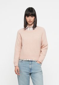 Maglione lavorato a maglia rosa chiaro con scollo rotondo e polsini a costine, abbinato a una camicia a righe con colletto sotto e jeans azzurri chiari.
