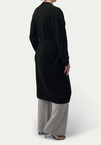 Lång svart kavaj med knytning i midjan, gjord av mjukt material; bärs över grå vida byxor. Har sänkta axlar och rak passform.