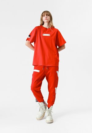 Camiseta roja de gran tamaño con una etiqueta rectangular blanca y acentos negros, combinada con pantalones joggers rojos que presentan diseños gráficos en blanco y cremalleras.