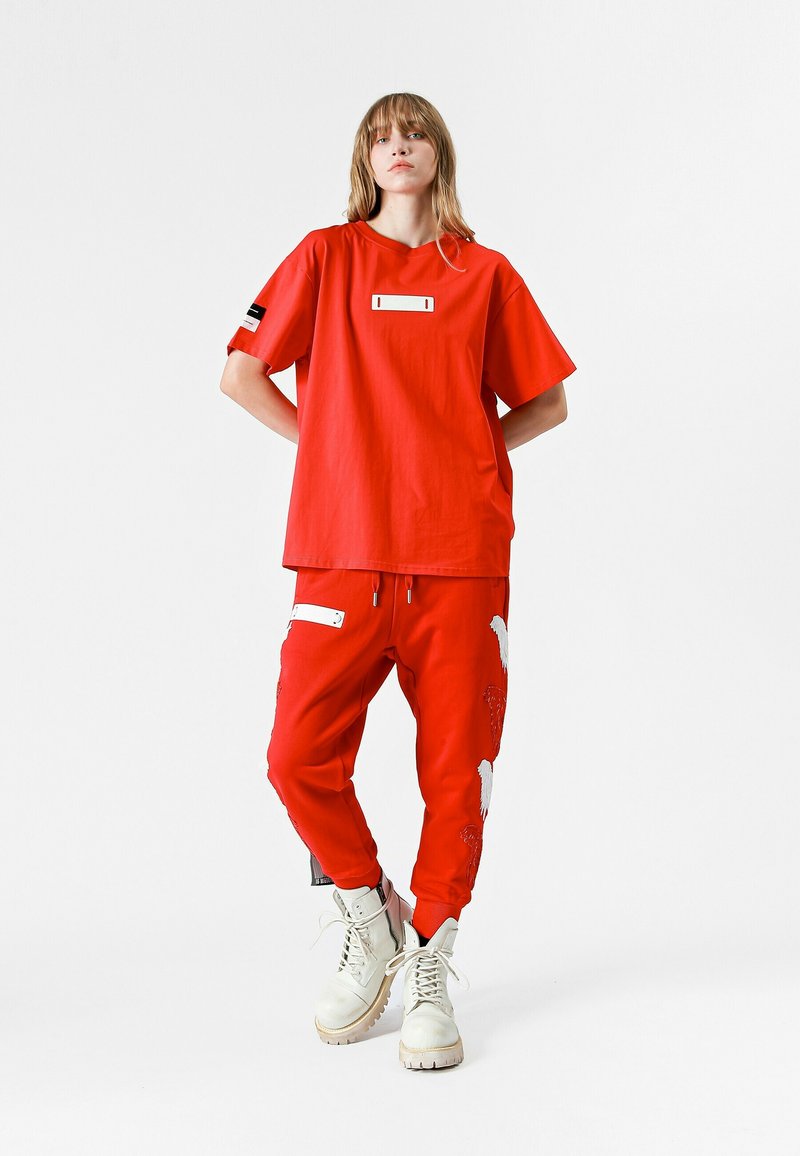Camiseta roja de gran tamaño con una etiqueta rectangular blanca y acentos negros, combinada con pantalones joggers rojos que presentan diseños gráficos en blanco y cremalleras.