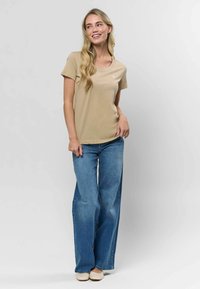 Béžové bavlnené tričko s okrúhlym výstrihom, skombinované s svetlomodrými širokými džínsami. Modelka stojí s jednou rukou na páse a má na sebe tkané topánky.