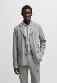 Lichtgrijs pak met een op maat gemaakte blazer met een notch lapel en twee knopen, gecombineerd met bijpassende broek en een lichtgrijze blouse eronder.
