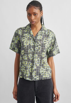 Tânără cu păr împletit, purtând o cămașă cu mâneci scurte, cu nasturi, cu model în carouri gri, galben și verde, combinat cu modele florale.