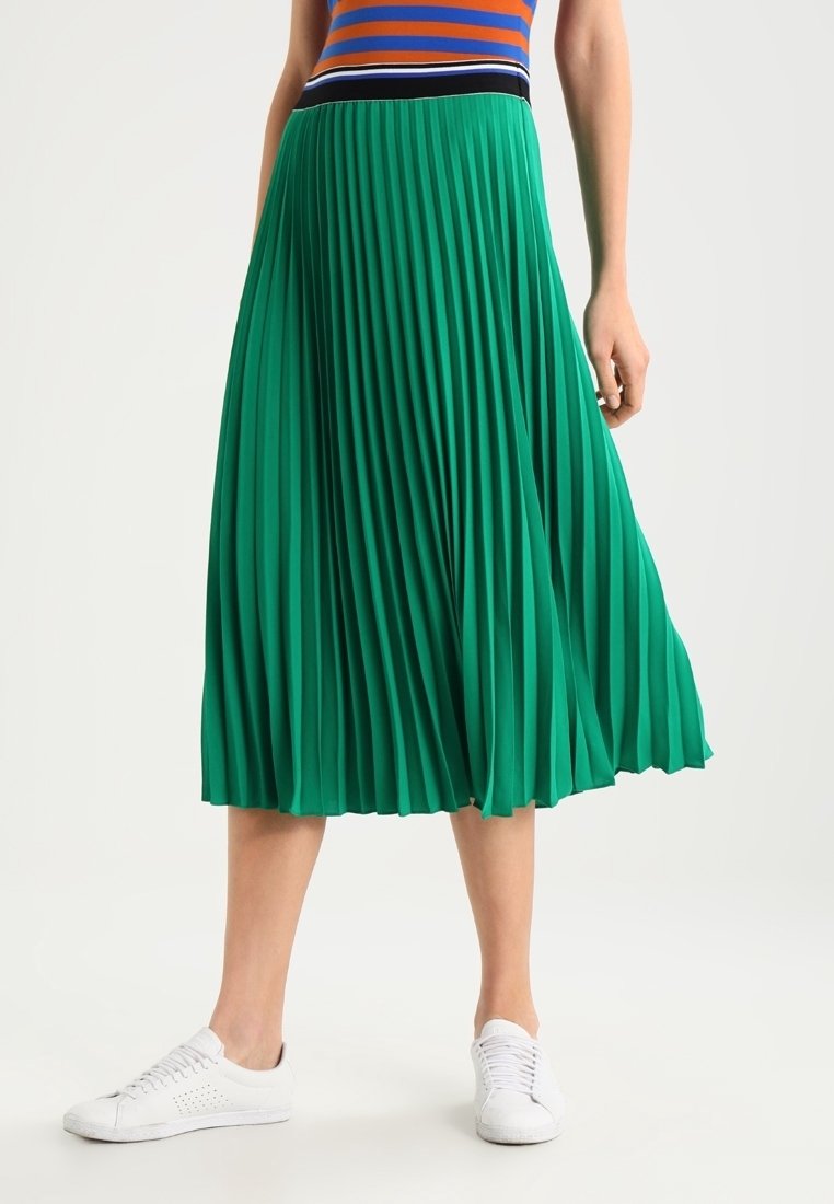 Falda midi plisada verde con una cintura negra y detalles blancos, combinada con zapatillas blancas. Textura de tela suave y diseño fluido.