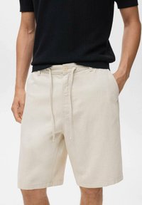 Pantalones cortos de algodón beige con cintura elástica y cordón, cierre de botón y bolsillos laterales. Tejido ligeramente texturizado, de longitud hasta la rodilla y con un ajuste relajado.