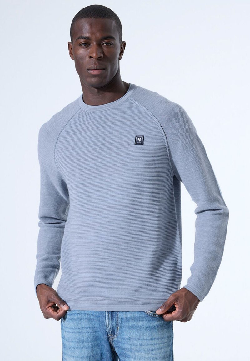 Suéter de manga larga gris claro con textura acanalada, cuello redondo y parche con el logo. Combinado con jeans de mezclilla azul, con las manos en la cintura.