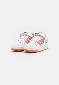 White athletic shoes with orange accents, featuring three stripes, a perforated toe, and a Velcro strap over laces. Rubber sole.