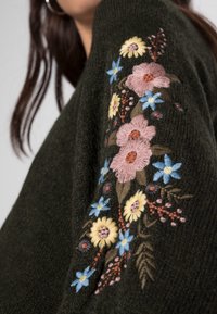 Un pull en maille vert foncé avec un brodé floral complexe en rose, jaune et bleu sur la manche, et une texture douce.