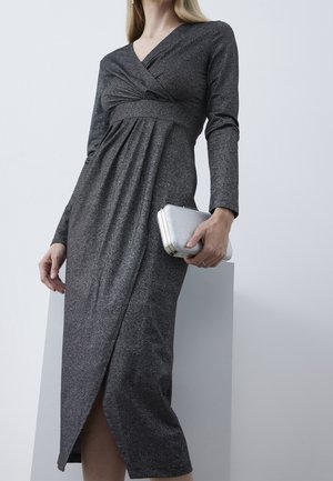 Kobieta w długim rękawie, ciemnoszarej sukience o kopertowym kroju, trzymająca srebrną kopertówkę na jasnym, jednolitym tle.