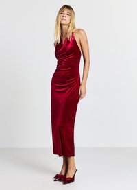 Abito in velluto rosso con scollo a halter e silhouette aderente, che arriva fino a metà polpaccio. Abbinato a tacchi a forma di fiocco rossi coordinati.