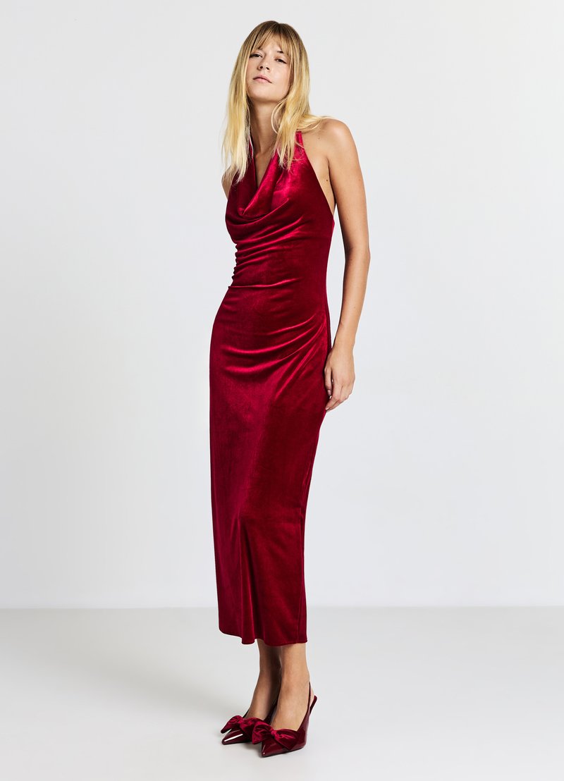 Abito in velluto rosso con scollo a halter e silhouette aderente, che arriva fino a metà polpaccio. Abbinato a tacchi a forma di fiocco rossi coordinati.