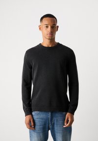 Schwarzer Strickpullover mit Rundhalsausschnitt, strukturiertem Stoff und gerippten Säumen, kombiniert mit hellblauen Jeans.