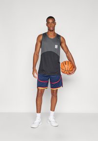 Sport tanktop i sort med en grå panel og logo, parret med marineblå shorts med rød og gul kant, og hvide sneakers. Holder en basketball.