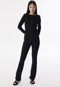 Schwarzes Langarmshirt mit Knopfleiste und strukturiertem Design, kombiniert mit schwarzen Slim-Fit-Hosen mit schrägen Nähten und verkürzten Säumen. Schwarze Sandalen.