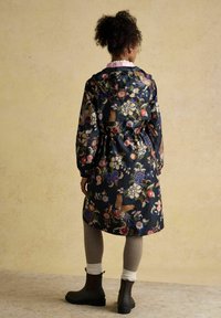 Femeie cu păr creț, purtând o pelerină de ploaie bleumarin cu model floral, colanți în carouri, șosete albe și ghete marron închis, stând cu spatele spre noi.