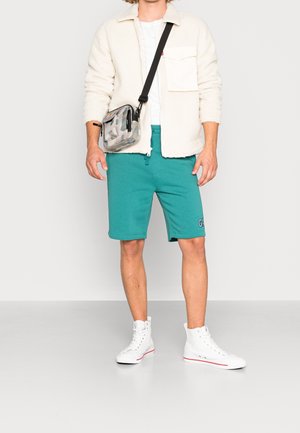 Veste en polaire beige clair avec poche à bouton, short teal et baskets montantes blanches. Sac en bandoulière transparent avec motif multicolore.