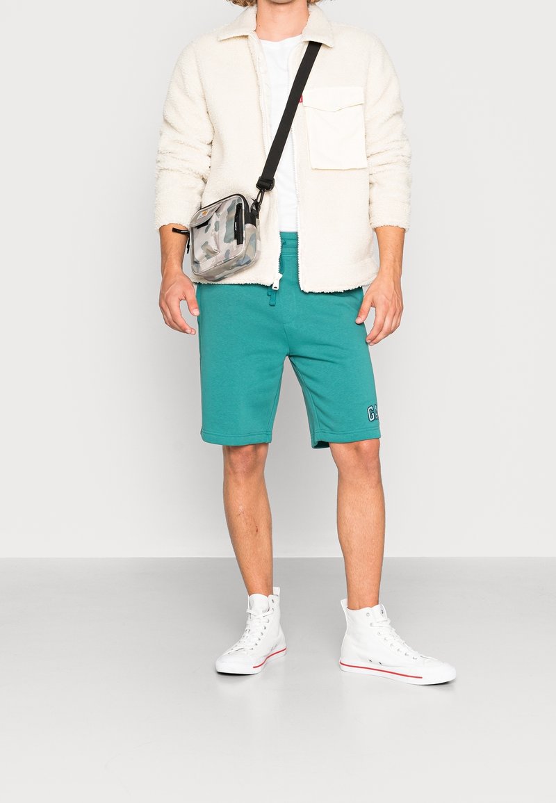 Giacca in pile beige chiaro con tasca a bottone, pantaloni corti teal e sneaker alte bianche. Borsa a tracolla trasparente con motivo multicolore.