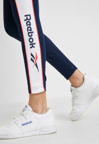 Ben i marinblå Reebok leggings med vita och röda ränder, och vita Reebok sneakers mot en enfärgad bakgrund.