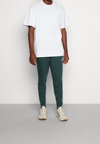 Muž v jednoduchém bílém tričku, tmavě zelených Adidas teplácích s bílými pruhy a béžových Adidas teniskách stojící na jednobarevném pozadí.