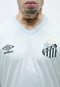 Balta sporta krekls ar gaiši zilu rakstainu v-veida kaklu, ar melnu logo un baltām svītrām, kā arī zīmola "Umbro" nosaukumu melnā krāsā.