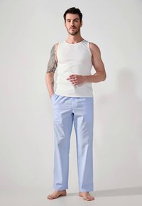 Man die blootsvoets staat in een wit mouwloos shirt en lichtblauwe gestreepte broek met een tattoo op zijn linkerarm.