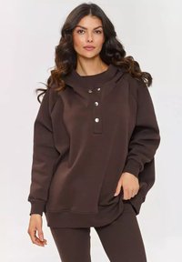 Brązowa lubiana bluza z kapturem, o luźnym kroju, z guzikami z przodu, ściągaczami przy rękawach i miękką fakturą. Połączona z pasującymi brązowymi legginsami.