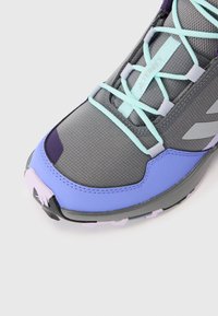 Chaussure de sport grise avec un dessus texturé, présentant des accents violets et bleu clair, ainsi qu'un design en maille. Inclut des lacets et une semelle durable.