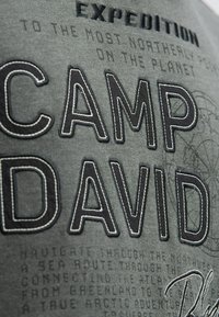 Graues Stoff-T-Shirt mit großem gesticktem Schriftzug "CAMP DAVID" in Schwarz, zusätzlichen Druckakzenten und einer strukturierten Oberfläche.