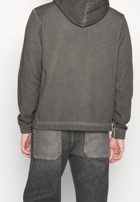 Felpa con cappuccio grigia e finitura testurizzata, mostrata da dietro. Abbinata a jeans di denim scuro con tasche applicate e cuciture a contrasto.