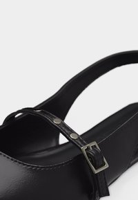 Μαύρο παπούτσι Mary Jane με λεπτό λουράκι που διαθέτει μεταλλικά καρφιά και ορθογώνια ασημί πόρπη, εμφανίζοντας τη φτέρνα και τις πλευρικές λεπτομέρειες.