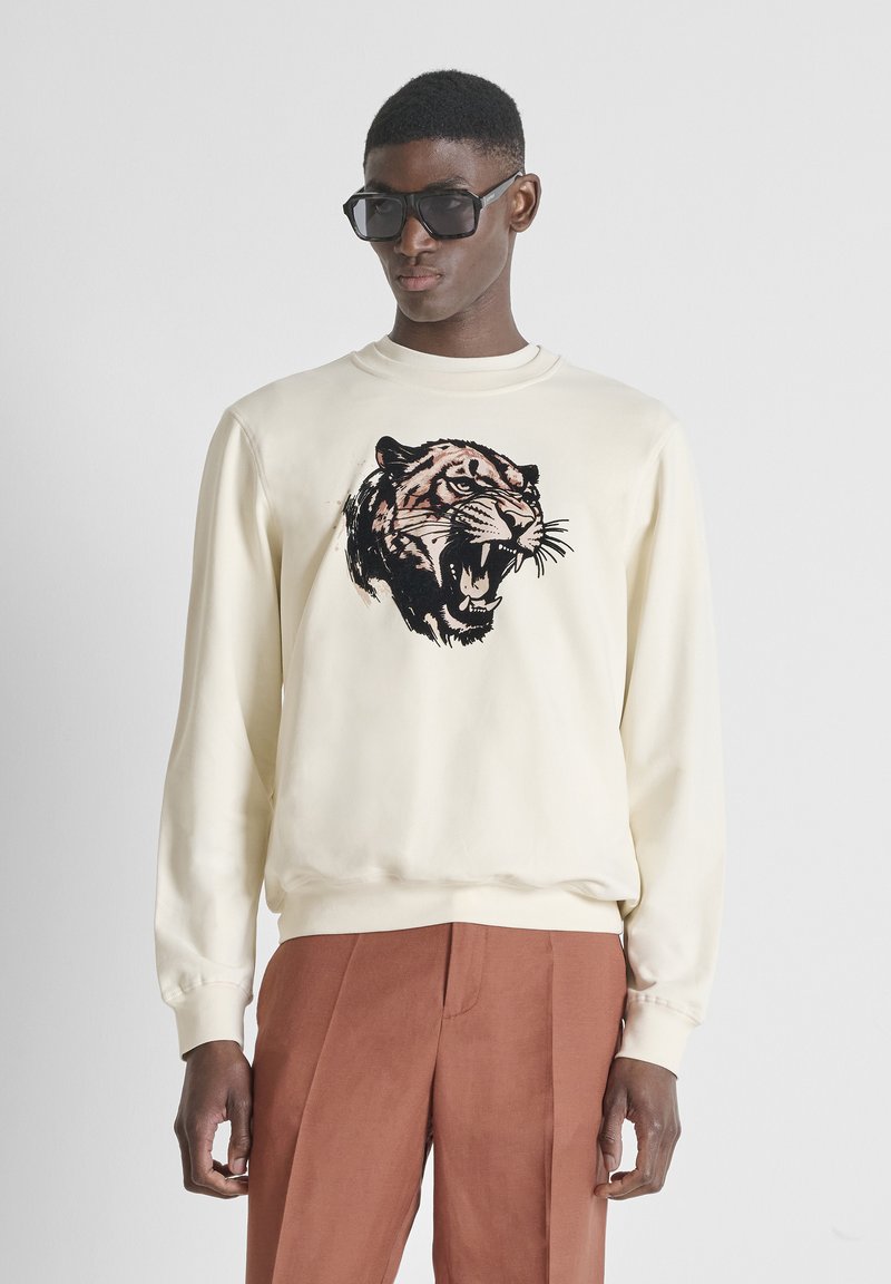 Hombre con gafas de sol negras de gran tamaño, sudadera color crema con gráfico de tigre rugiente y pantalones color óxido, de pie frente a un fondo liso.