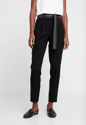 Mujer con pantalones negros ajustados con dobladillos con puños y un cinturón negro con rayas blancas, combinado con zapatos negros sin cordones.